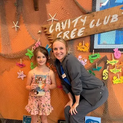A young Cavity Free Club winner with her photo prize and a Super Smiles team member, in front of the Cavity Free Club wall — fishing-net display with the hand-painted "CAVITY FREE CLUB" sign
